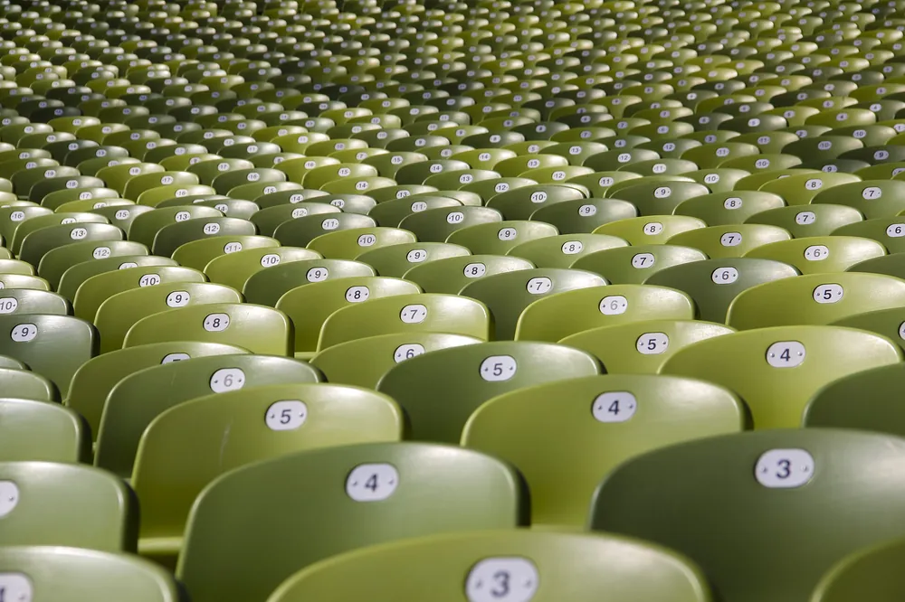 Filas de cadeiras verdes numeradas numa bancada vazia de um pavilhão