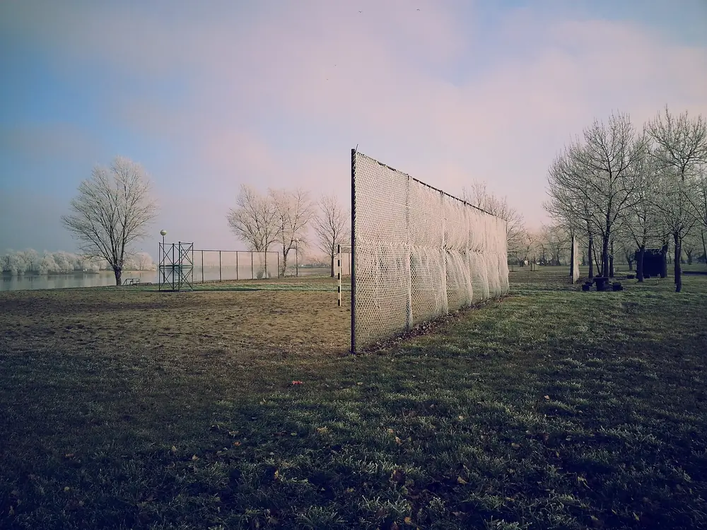 Campo de treino ao ar livre com rede de proteção alta e árvores despidas ao fundo